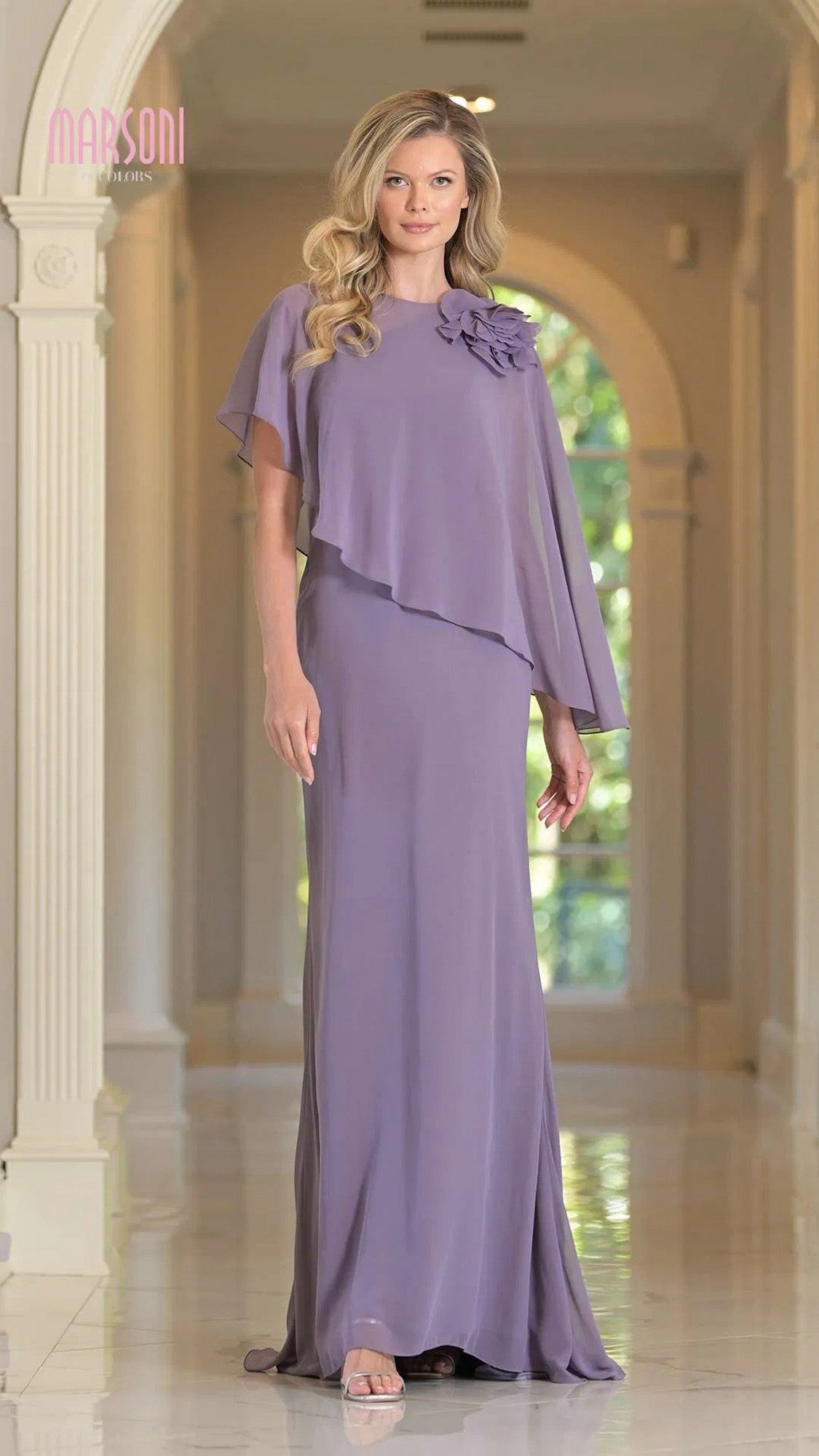 A woman stands in a hallway wearing the Marsoni MV1347 long formal dress in wisteria, featuring floor-length chiffon, a layered cape, and floral shoulder detail.